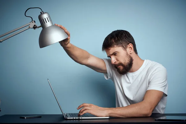 Un hombre con un portátil en el interior en una mesa sobre un fondo azul y lámpara oficina de trabajo — Foto de Stock