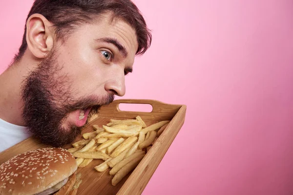 Muž s tácem s jídlem v ruce hamburger hranolky a rychlé občerstvení kalorie růžové pozadí portrét close-up — Stock fotografie