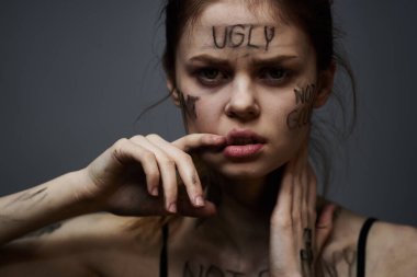 Hüsrana uğramış bir kadın vücudunda çirkin yazıtlar ve gri bir arka plan portresi üzerinde siyah bir tişört var. 