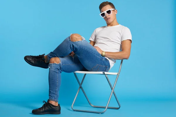 Ragazzo rilassato in jeans con le ginocchia strappate si siede su una sedia al coperto su uno sfondo blu con occhiali a figura intera sul suo viso — Foto Stock