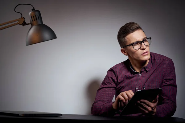 Hombre de negocios con gafas y una camisa sentada en una mesa en una habitación oscura con una lámpara vista recortada — Foto de Stock