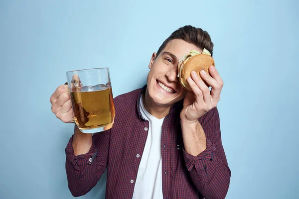 Alegre bêbado homem com caneca de cerveja e hambúrguer na mão dieta comida estilo de vida azul fundo — Fotografia de Stock