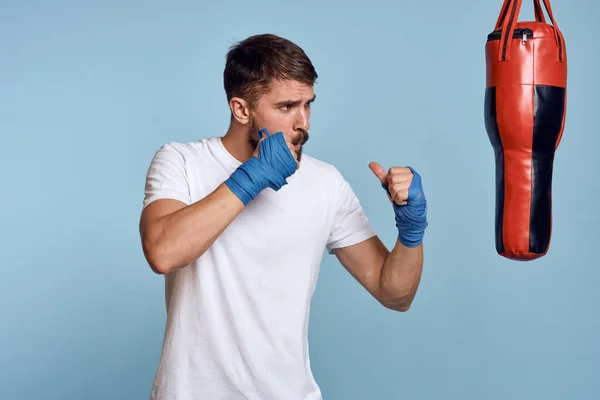 Un hombre practicando un puñetazo en un saco de boxeo en una camiseta blanca sobre un fondo azul — Foto de Stock