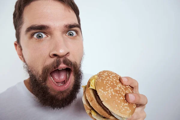 Ein Mann, der einen Hamburger auf hellem Hintergrund in einem weißen T-Shirt isst, hat die Nahaufnahme von Hunger-Fast Food abgeschnitten — Stockfoto