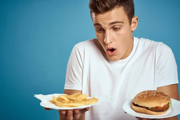 Jovem com batatas fritas e hambúrguer no fundo azul interessado olhar emoções fast food calorias corte vista Copiar Espaço — Fotografia de Stock