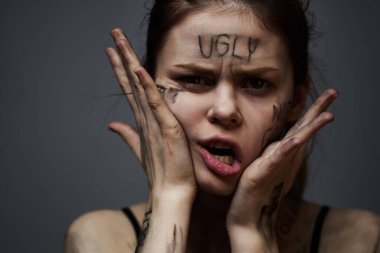 Gri arka planda kırıcı yazılar yazan üzgün kadın elleriyle kendine dokunuyor.