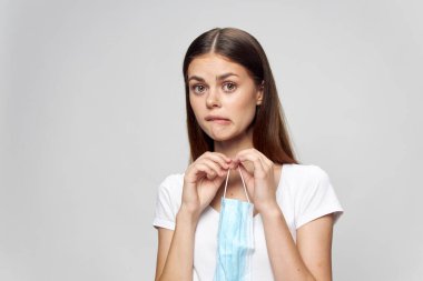 Elinde tıbbi maske olan bir kadın ileriye bakıyor dudak beyazı tişörtünü ısırıyor. 