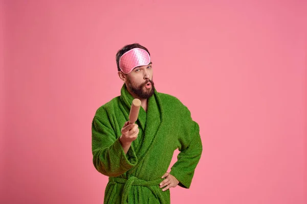 Retrato de um homem em uma máscara de sono rosa e um rolo de madeira emoções modelo de irritabilidade roupão verde — Fotografia de Stock