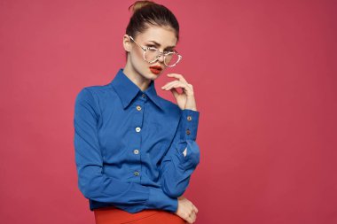 Gözlüklü ve mavi gömlekli bir kadının portresi. Kırmızı dudaklı, pembe arka plan modeli.