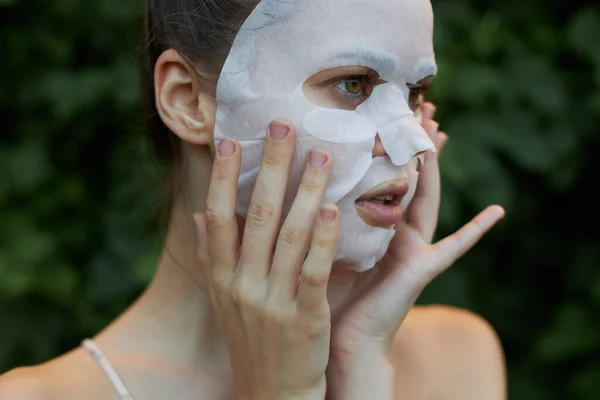 Mujer hermosa máscara blanca Mira a un lado y mantener las manos cerca de su cara arbustos de cosmetología en el fondo —  Fotos de Stock