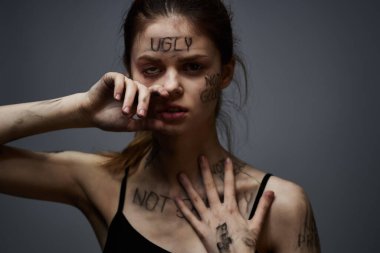 Hüsrana uğramış bir kadın vücudunda çirkin yazıtlar ve gri bir arka plan portresi üzerinde siyah bir tişört var. 