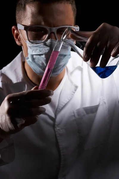 Assistant de laboratoire dans un masque médical et dans un manteau blanc avec un liquide dans une fiole et un produit chimique sur un fond noir — Photo