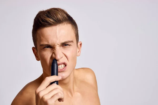 Ragazzo con un trimmer in mano rimuovere i capelli dal naso spalle nude emozioni sfondo chiaro — Foto Stock