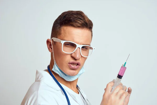 Medico con uno stetoscopio in occhiali tiene una siringa tra le mani su uno sfondo chiaro vista laterale — Foto Stock