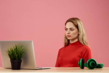 Dizüstü bilgisayarın önündeki masada oturan bir kadın elinde bir oyun platformuyla teknoloji kulaklığı çalıyor. 