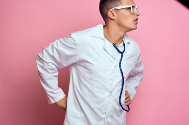 Pembe bir arka planda steteskop ve yüzünde gözlük olan tıbbi önlüklü bir erkek doktor.