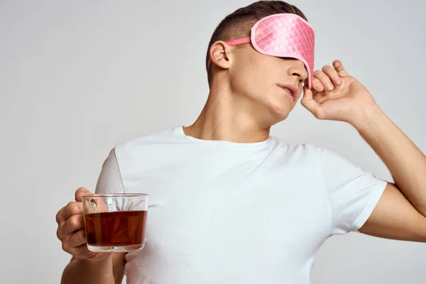 Homem bonito com máscara de sono rosa e com uma xícara de chá na mão puxa-se sobre um fundo leve — Fotografia de Stock