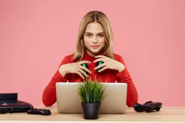 Kulaklık takan, dizüstü bilgisayarlı bir kadın masa başı eğlence sektöründe pembe arka planda oturuyor.