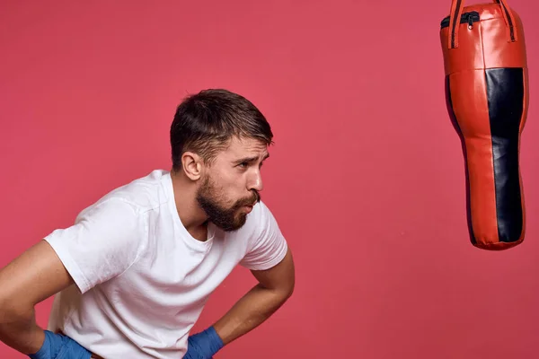 Un homme près d'un sac de boxe en gants bleus et un T-shirt blanc pratique des coups de poing sportifs — Photo