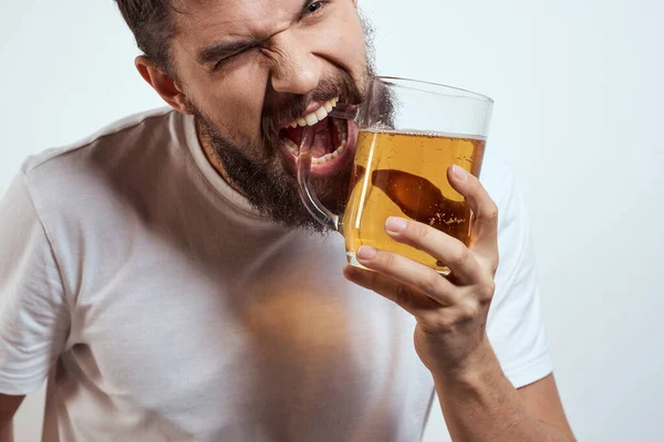 Um homem barbudo com uma caneca de cerveja em um fundo leve em uma camiseta branca vista recortada de uma bebida alcoólica — Fotografia de Stock