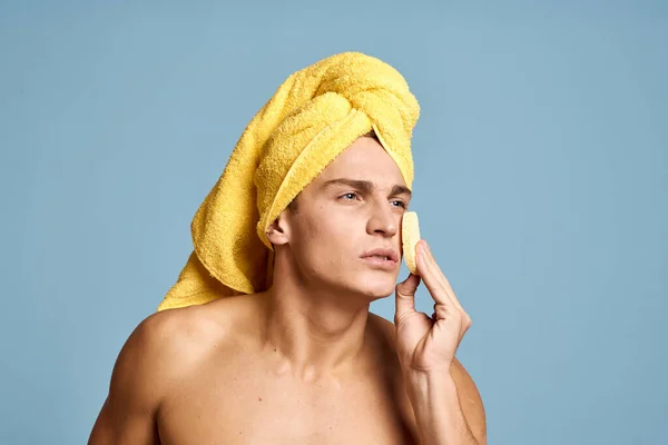 Un uomo con un asciugamano giallo sulla testa e una spugna morbida per la pulizia della pelle modello cosmetologia — Foto Stock