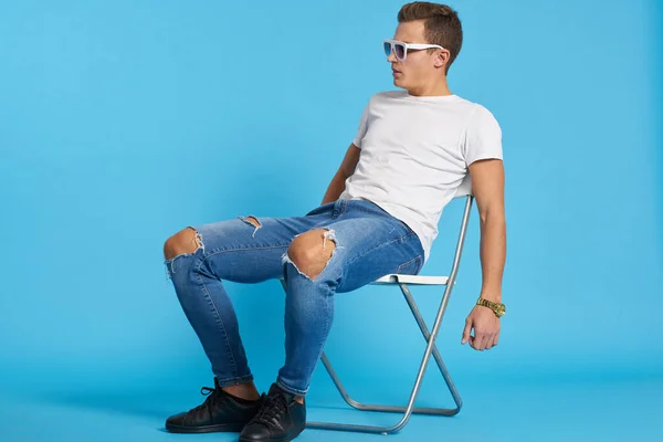 Ragazzo rilassato in jeans con le ginocchia strappate si siede su una sedia al coperto su uno sfondo blu con occhiali a figura intera sul suo viso — Foto Stock