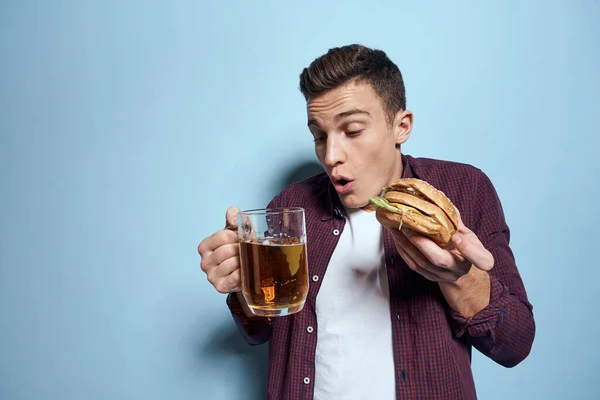 Alegre bêbado homem com caneca de cerveja e hambúrguer na mão dieta comida estilo de vida azul fundo — Fotografia de Stock