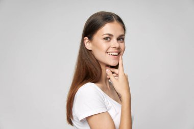 Beyaz tişörtlü bir kadın gülümsüyor. Parmağını ağzının yakınında tutuyor. Uzun saçlı. 