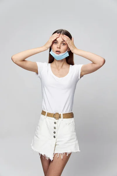 Femme surprise dans un T-shirt blanc avec un masque médical sur le visage touche sa tête avec ses mains — Photo
