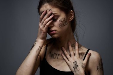 Hüsrana uğramış bir kadın vücudunda çirkin yazıtlar ve gri bir arka plan portresi üzerinde siyah bir tişört var. 
