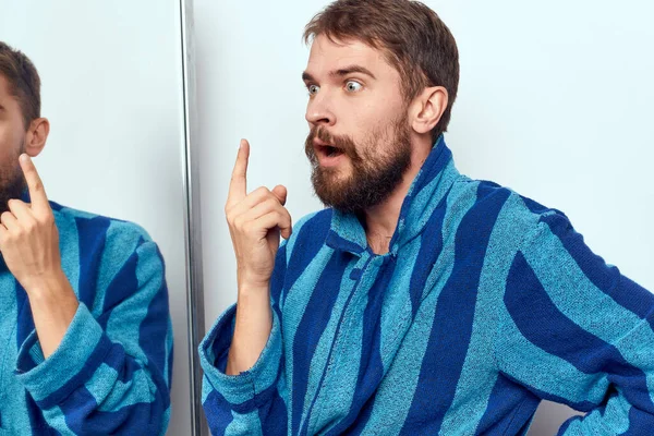 Человек в синем халате рассматривает себя в зеркале в яркой комнате обрезанный вид — стоковое фото