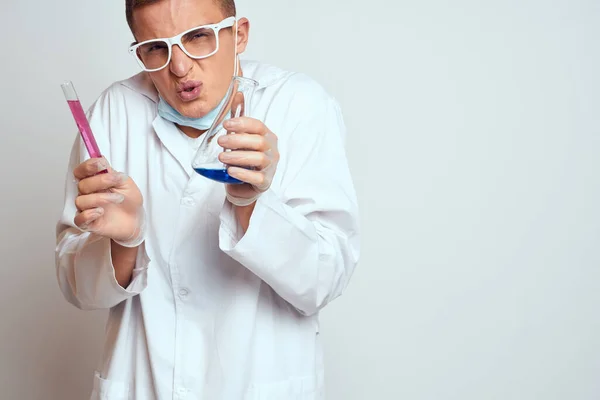 Un assistant de laboratoire en blouse médicale avec un masque de protection tient une fiole avec un liquide de réaction chimique — Photo