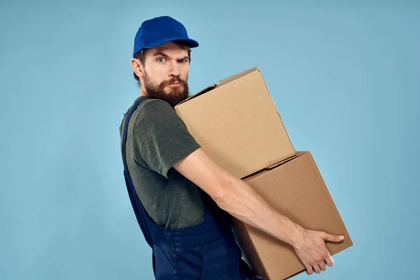 Muž v pracovní uniformě s krabicemi v ruce dodávka služby modré pozadí — Stock fotografie