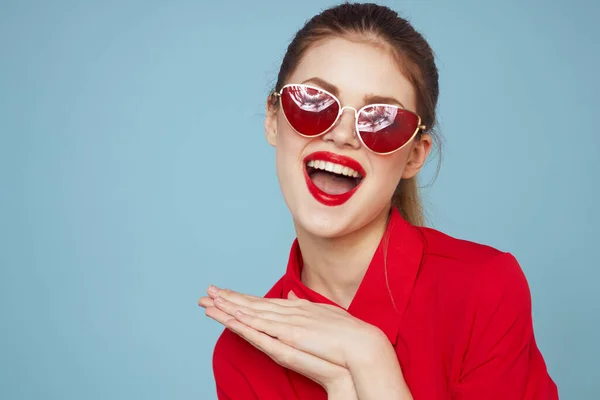 Mujer con camisa roja sobre fondo azul sostiene las manos cerca de la cara y gafas de sol labios rojos modelo de maquillaje — Foto de Stock