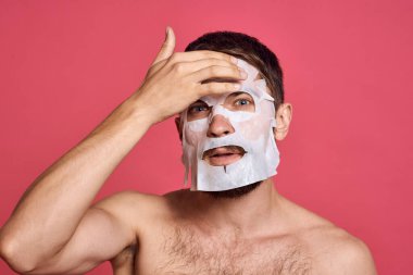 Pembe arka planda maske takan, elleri çıplak, gövdesi kesilmiş bir adam.