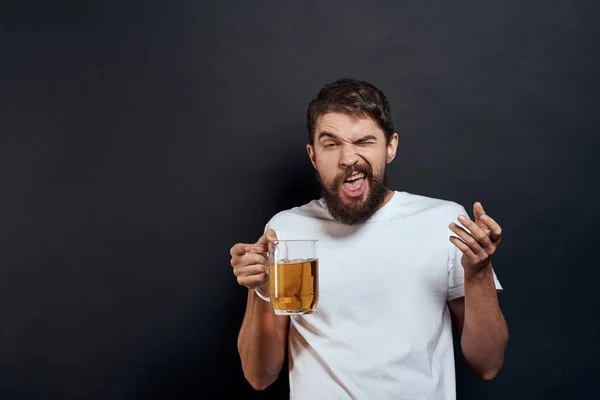Elinde bir kupa birayla gezen bir adam duyguları olan eğlenceli bir yaşam tarzı beyaz tişört koyu tenha bir arka plan. — Stok fotoğraf