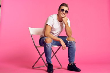 Beyaz tişörtlü ve kot pantolonlu bir adam, modern tarz pembe izole edilmiş arka planda oturuyor.