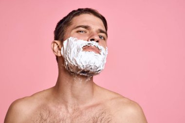 Yüzünde jilet ve beyaz köpük olan bir adam. Pembe arka planda çıplak gövde banyo tedavileri.