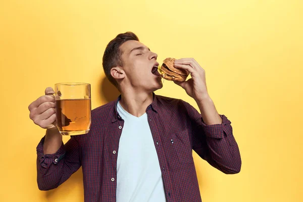 Cheerful man with a mug of beer and a hamburger on his hands diet food lifestyle yellow background — Stock Photo, Image