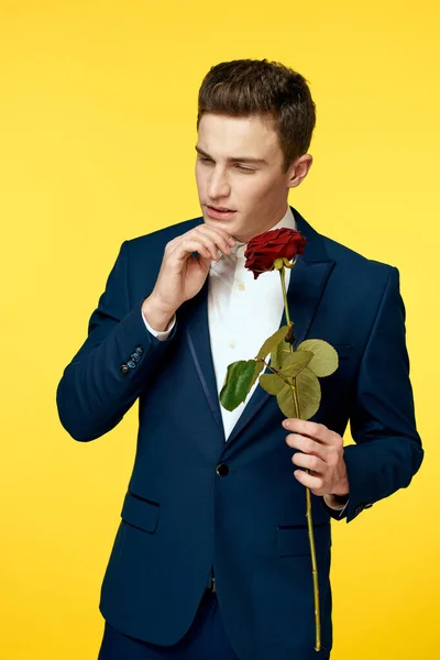 Junger Mann im klassischen Anzug mit roter Rose in der Hand auf gelbem Hintergrund — Stockfoto