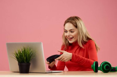 Dizüstü bilgisayarın önündeki masada oturan bir kadın elinde bir oyun platformuyla teknoloji kulaklığı çalıyor. 