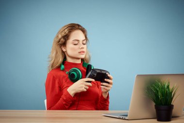 Dizüstü bilgisayar kulaklığının önündeki masada oturan bir kadın çevrimiçi yaşam tarzı mavi arka planda oynuyor.