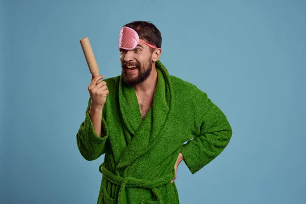 A man with a pink sleep mask holds a rolling pin in his hand and a green robe blue background emotions model — Stock Photo, Image