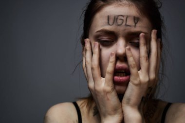 Gri arka planda kırıcı yazılar yazan üzgün kadın elleriyle kendine dokunuyor.