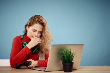 Dizüstü bilgisayar kulaklığının önündeki masada oturan bir kadın çevrimiçi yaşam tarzı mavi arka planda oynuyor.