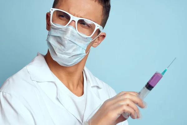 Dokter memegang jarum suntik di tangannya dengan cairan merah pada latar belakang biru gaun medis dan topeng pelindung — Stok Foto