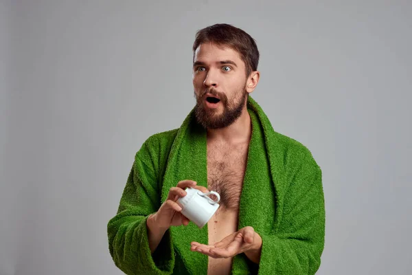 Homem em um manto verde com uma xícara de bebida em suas mãos em um fundo cinza vista cortada — Fotografia de Stock