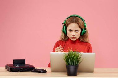 Kulaklık takan, dizüstü bilgisayarlı bir kadın masa başı eğlence sektöründe pembe arka planda oturuyor.