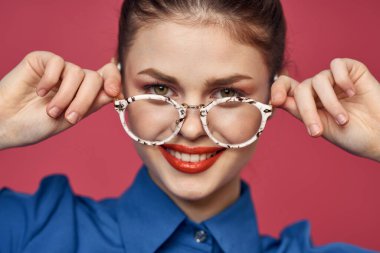 Gözlüklü ve mavi gömlekli bir kadının portresi. Kırmızı dudaklı, pembe arka plan modeli.