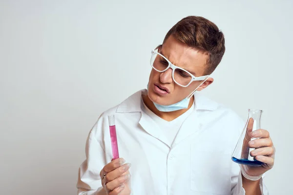 Un assistant de laboratoire en blouse médicale avec un masque de protection tient une fiole avec un liquide de réaction chimique — Photo